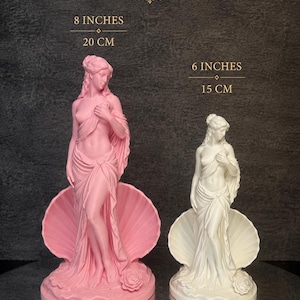 May include: Two statues of a figure standing in a seashell, one pink and one white. The pink statue is 20 cm (8 inches) tall, and the white statue is 15 cm (6 inches) tall. Both figures have draped fabric, detailed hair, and a rose at the base.