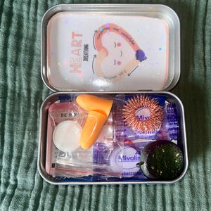 Könnte beinhalten: Eine offene, rechteckige Metallbox mit Deckel, auf dem die Worte "HEART BREATHING" und eine herzförmige Illustration stehen. Im Inneren befinden sich verschiedene Gegenstände, darunter Ohrstöpsel, ein stacheliger Massagering und kleine Päckchen. Die Box steht auf einer grünen Oberfläche.