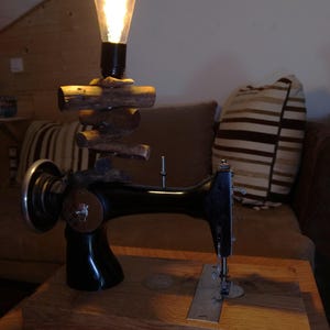 Könnte beinhalten: Eine schwarze Vintage-Nähmaschinenbasis auf einem rechteckigen Holzblock, mit einem Stapel kleiner horizontaler Holzscheite, gekrönt von einer leuchtenden Edison-Glühbirne. Die Metallmaschine verfügt über ein silbernes Handrad.