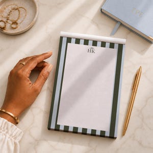 May include: A rectangular notepad featuring a vertical striped border in dark green and light blue. The center of the notepad is white with the monogram HK printed in black at the top. A gold pen is positioned to the right of the notepad.