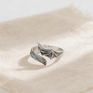 Op de afbeelding: Een zilverkleurige ring met een drakenvleugelontwerp. De ring heeft gedetailleerde vleugeltexturen en een gesplitste band voor een verstelbare pasvorm. De ring wordt getoond op een beige stoffen oppervlak.