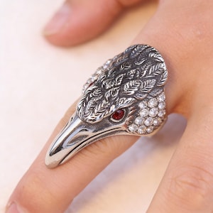 Puede incluir: Un anillo plateado con forma de cabeza de p&aacute;jaro, con un ojo de gema roja y cristales transparentes. La cabeza del p&aacute;jaro tiene grabados detallados de plumas. El anillo se lleva en un dedo.