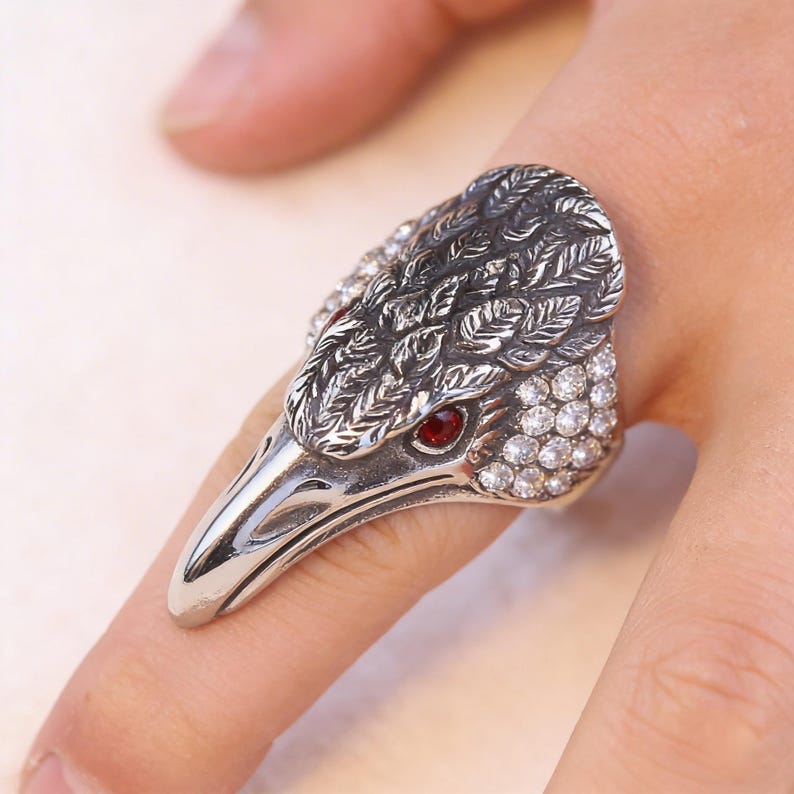 Puede incluir: Un anillo plateado con forma de cabeza de p&aacute;jaro, con un ojo de gema roja y cristales transparentes. La cabeza del p&aacute;jaro tiene grabados detallados de plumas. El anillo se lleva en un dedo.