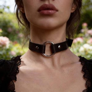 Op de afbeelding: Een zwarte leren choker ketting met een grote zilveren ring in het midden. De choker is vastgemaakt met een gesp en twee zilveren klinknagels. De achtergrond is wazig, met hints van groen en roze.