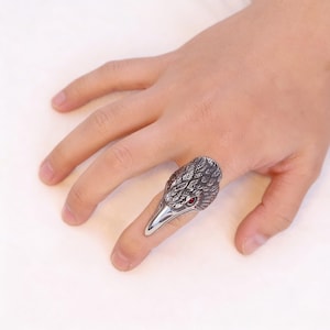 Puede incluir: Anillo plateado con forma de cabeza de p&aacute;jaro, con detalles de plumas y un ojo de gema roja. El anillo se lleva en un dedo, mostrando su dise&ntilde;o &uacute;nico inspirado en las aves.