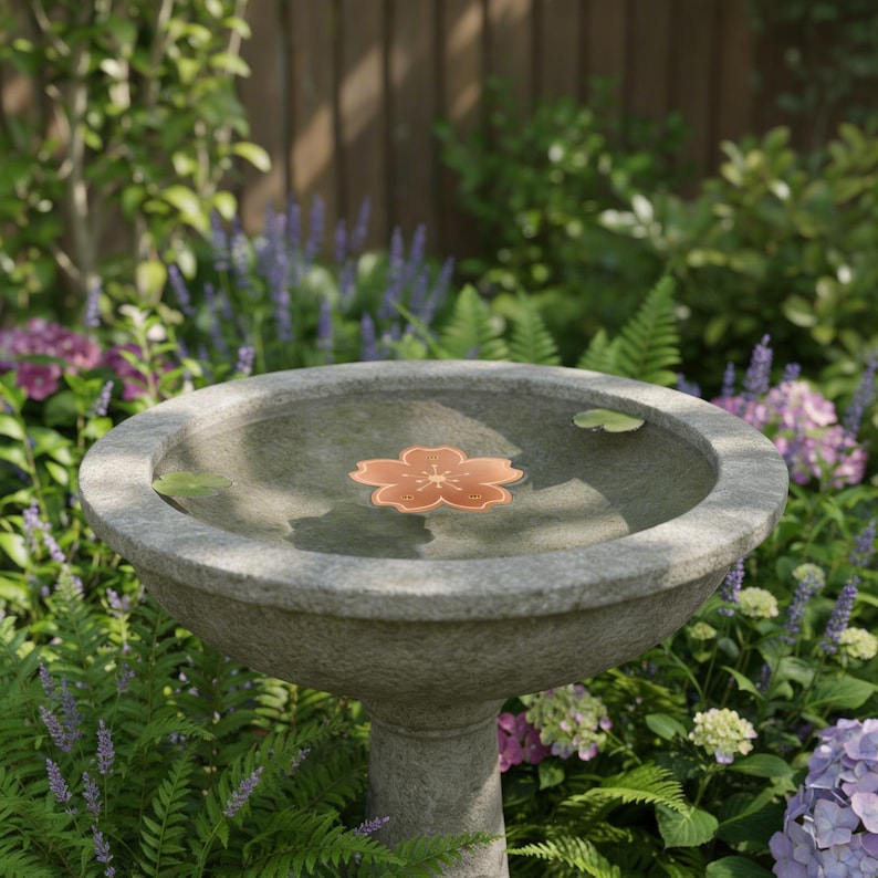 Puede incluir: Un bebedero de piedra gris con una decoraci&oacute;n flotante en forma de flor de color cobre. El bebedero est&aacute; rodeado de exuberante vegetaci&oacute;n, incluyendo helechos, flores moradas y hortensias, con una valla de madera al fondo.