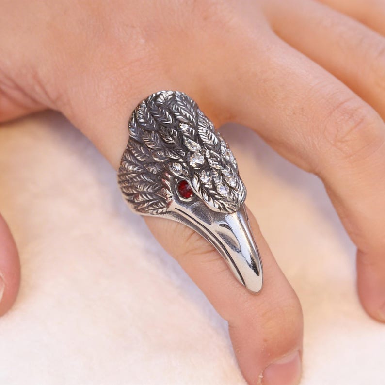 Puede incluir: Anillo plateado con forma de cabeza de p&aacute;jaro, con un dise&ntilde;o detallado de plumas y un ojo de gema roja. El anillo se lleva en un dedo, mostrando la intrincada artesan&iacute;a y el estilo &uacute;nico. Una joya llamativa.