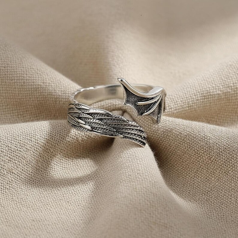 Op de afbeelding: Een zilverkleurige ring met een vleugel- en vleermuisvleugelontwerp. De ring heeft gedetailleerde veer- en vleugeltexturen. De ring is geplaatst tegen een neutrale stoffen achtergrond.