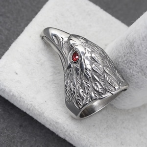 Puede incluir: Anillo de plata con forma de cabeza de &aacute;guila, con detalles de plumas y un ojo de gema roja. El anillo se muestra sobre un fondo blanco, resaltando el dise&ntilde;o intrincado y la artesan&iacute;a.