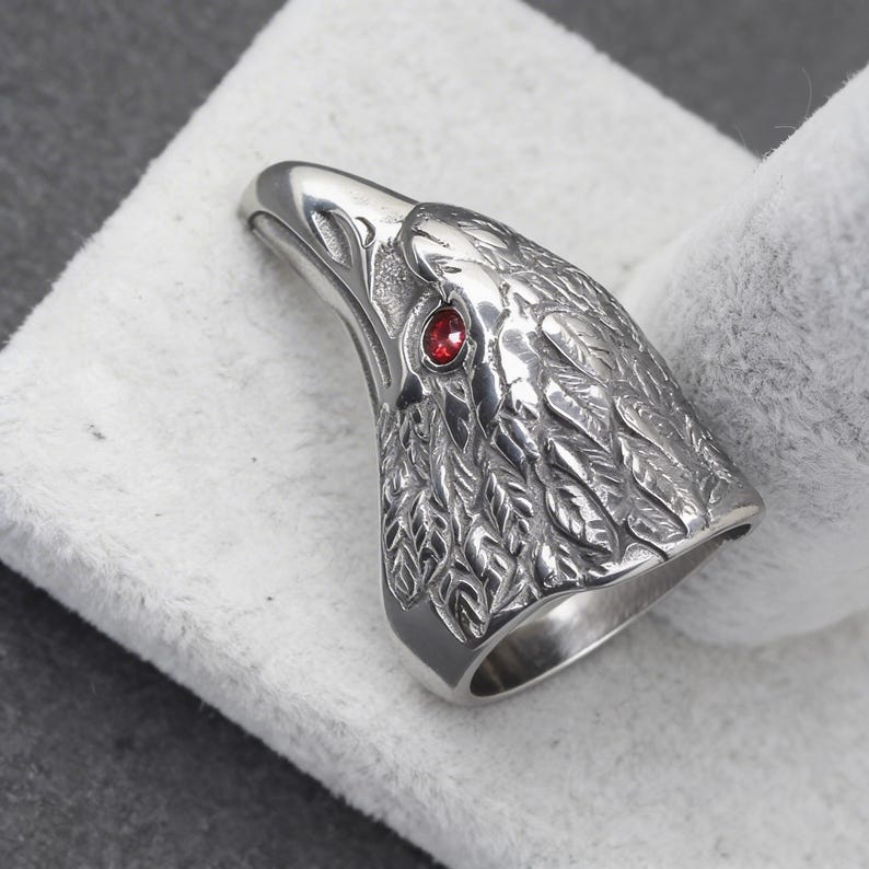 Puede incluir: Anillo de plata con forma de cabeza de &aacute;guila, con detalles de plumas y un ojo de gema roja. El anillo se muestra sobre un fondo blanco, resaltando el dise&ntilde;o intrincado y la artesan&iacute;a.