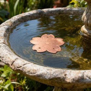 Op de afbeelding: Een koperkleurige, bloemvormige fontein drijver rust in een stenen fontein gevuld met water. De drijver heeft een gedetailleerd bloemblaadjesontwerp en kleine gaatjes. De fontein bevindt zich buiten, omringd door groen.