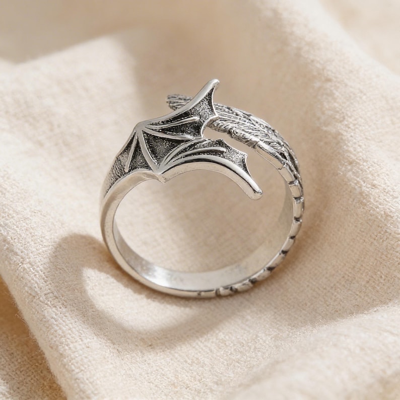 Op de afbeelding: Een zilverkleurige ring met een drakenvleugelontwerp. De ring heeft een gedetailleerde vleugelvorm met getextureerde lijnen en een gebogen band. De ring is geplaatst tegen een neutrale stoffen achtergrond.