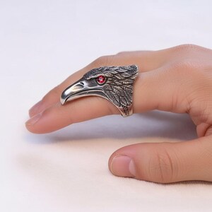 Puede incluir: Un anillo plateado con forma de cabeza de &aacute;guila, con un ojo de piedra roja. El anillo, que se lleva en un dedo, presenta detalles de plumas. El pico del &aacute;guila est&aacute; abierto y el anillo tiene una est&eacute;tica vintage o antigua.