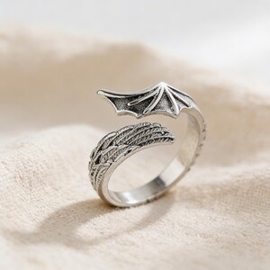 Op de afbeelding: Een zilverkleurige ring met een drakenvleugelontwerp. De ring heeft een gedetailleerde vleugel aan de ene kant en een gestileerde vleugel aan de andere kant, waardoor een open, verstelbaar ontwerp ontstaat. De ring staat tegen een neutrale achtergrond.