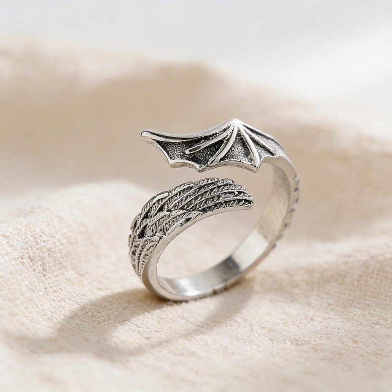 Op de afbeelding: Een zilverkleurige ring met een drakenvleugelontwerp. De ring heeft een gedetailleerde vleugel aan de ene kant en een gestileerde vleugel aan de andere kant, waardoor een open, verstelbaar ontwerp ontstaat. De ring staat tegen een neutrale achtergrond.