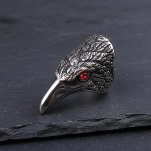 May include: A silver-colored ring shaped like a raven's head. The ring features detailed feather textures and a red gemstone eye. The raven's beak is pointed, and the ring is set against a dark background.