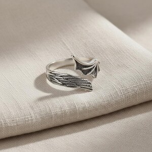 Op de afbeelding: Een zilverkleurige ring met een drakenvleugelontwerp. De ring heeft gedetailleerde veer- en vleugelpatronen, met een getextureerde afwerking. De ring staat tegen een neutrale achtergrond, waardoor de ingewikkelde details van de sieraden worden benadrukt.
