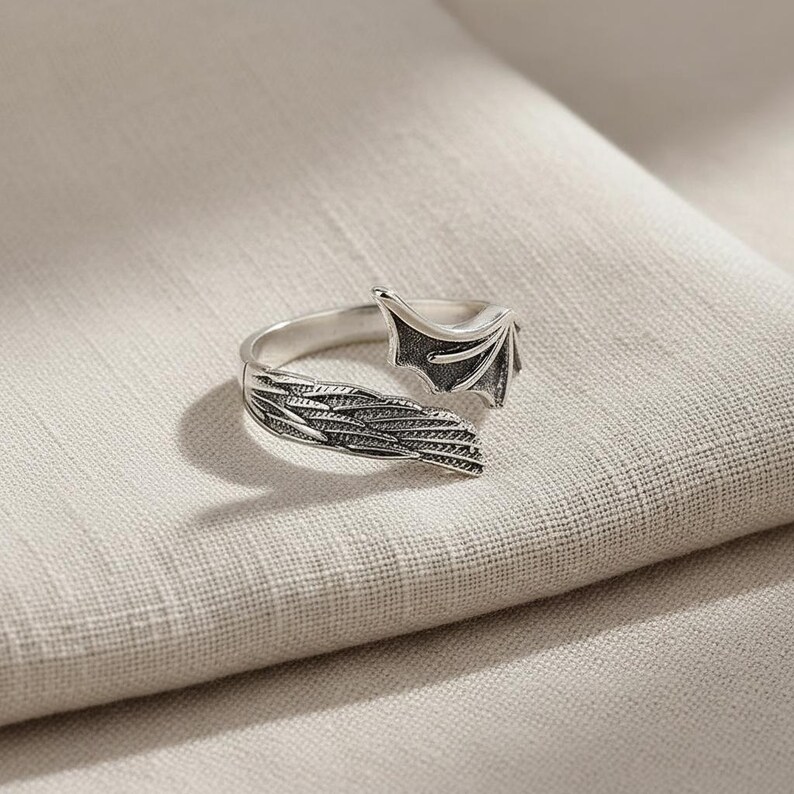 Op de afbeelding: Een zilverkleurige ring met een drakenvleugelontwerp. De ring heeft gedetailleerde veer- en vleugelpatronen, met een getextureerde afwerking. De ring staat tegen een neutrale achtergrond, waardoor de ingewikkelde details van de sieraden worden benadrukt.