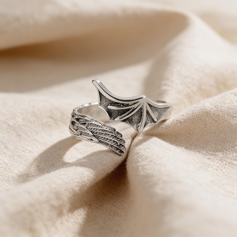 Op de afbeelding: Zilverkleurige ring met een drakenvleugelontwerp. De ring heeft aan elke kant een gedetailleerde vleugel, die zich om de vinger wikkelt. De vleugels zijn ingewikkeld gedetailleerd. De ring staat tegen een neutrale stoffen achtergrond.