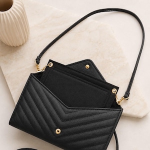 May include: A black envelope-style crossbody bag with a chevron-quilted texture, featuring a gold-tone snap button closure, a thin matching shoulder strap, and an open flap revealing a black interior compartment with a gold-tone snap detail.
