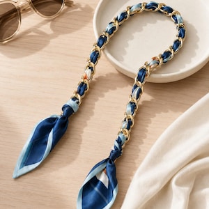 May include: A decorative chain strap featuring a gold-tone metal link design interwoven with a smooth, navy blue and light blue patterned fabric ribbon. The strap ends in two loose, pointed fabric tails with a geometric print in shades of blue.