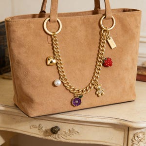 May include: A tan suede tote bag with gold-colored handles and a decorative chain. The chain features charms including a heart, pearl, flower, strawberry, bear, and a small tag. The bag is sitting on a white table.