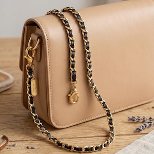 Op de afbeelding: Een beige leren handtas met een gouden kettingriem met zwart leer door de schakels geweven. De riem is met gouden sluitingen aan de tas bevestigd. Een klein goudkleurig label leest "HANDCRAFTED EST. 2023".