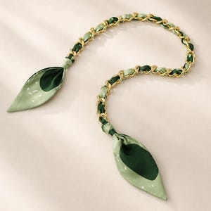 May include: A decorative bag strap featuring a gold-tone metal chain interwoven with a light green and dark green patterned fabric. The strap ends in two pointed fabric tabs with a dark green circular design and small white speckles.