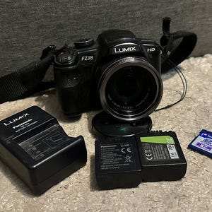 May include: Black Lumix FZ38 digital camera with a lens, strap, charger, two batteries, and an SD card. The camera has the Lumix and HD logos. The SD card is blue with white text. The charger is black and has the Lumix logo.