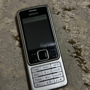 May include: A silver Nokia cell phone with a black screen and keypad. The phone displays the date 01/01/2008 and time 01:16pm. The keypad has white numbers and letters. The phone is on a textured surface.