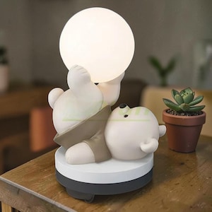 May include: A whimsical white bear-shaped lamp with a round, glowing light. The bear is lying down, holding the light above its head. The lamp has a brown vest and sits on a white and gray base. A small succulent plant in a brown pot is in the background.