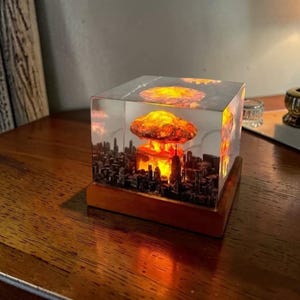 May include: A clear resin cube featuring a miniature cityscape with a mushroom cloud explosion. The explosion is illuminated with orange and yellow light, set against a gray sky. The cube sits on a wooden base.