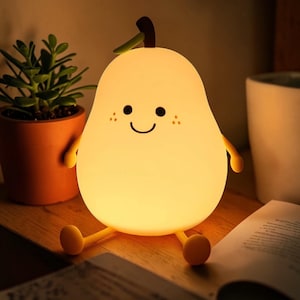 Pode incluir: Uma luz noturna alegre em forma de pêra, com um rosto sorridente, sardas e pequenos braços e pernas. A luz é de cor amarelo suave, com um caule verde e um topo castanho. A lâmpada está numa superfície de madeira.