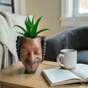 Może przedstawiać: Brązowa i czarna doniczka w kształcie twarzy z uśmiechniętą miną, trzymająca zieloną roślinę sukulentową. Doniczka stoi na drewnianym stole obok białego kubka i otwartej książki. Doniczka ma czarne włosy.