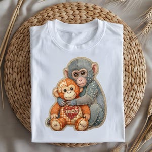 Puede incluir: Camiseta blanca con una colorida ilustración de dos monos abrazándose. Un mono es naranja con un corazón que dice "TEAM", el otro es gris. Las palabras "BE KIND" también están en el diseño.