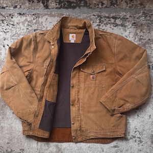 May include: A brown work jacket with a button-up front, two chest pockets, and a collar. The jacket has a brown interior lining and a small logo patch on the chest pocket. The jacket is made of a durable material.