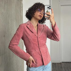 Op de afbeelding: Een stoffige roze blazer met een revers, twee voorzakken en een sluiting met één knoop. De jas wordt gecombineerd met een lichtblauwe jeans. De blazer lijkt gemaakt van suède of een vergelijkbaar zacht materiaal.