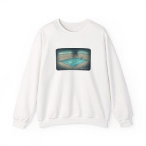 Könnte beinhalten: Weißes Sweatshirt mit Rundhalsausschnitt und rechteckiger Grafik eines Schwimmbeckens. Das Becken ist mit blauem Wasser gefüllt und hat eine geflieste Umgebung. Die Grafik ist in einer dunklen, gedämpften Farbpalette gehalten.