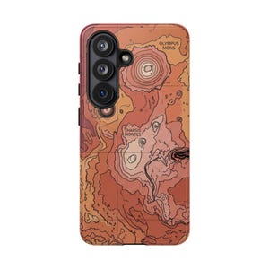 May include: A phone case featuring a topographic map design in shades of orange and brown. The case includes the text "OLYMPUS MONS" and "THARSIS MONTES". The case has a camera module with three lenses.