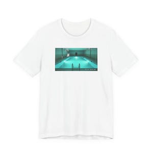 Könnte beinhalten: Weißes T-Shirt mit einem rechteckigen Bild eines Hallenbades mit türkisfarbenem Wasser. Das Becken hat eine Leiter und ist von einem gefliesten Bereich umgeben. Das Bild hat Text unten.