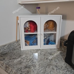 May include: A white cabinet with two arched glass doors, containing food items. The cabinet is on a countertop with a speckled pattern. A small container with a purple lid sits on top of the cabinet.