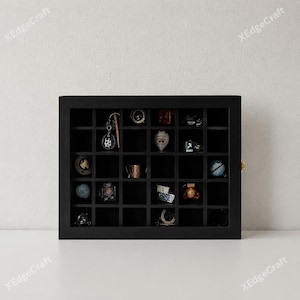 May include: A black display case with multiple compartments, showcasing a collection of small items. The case is rectangular with a grid of square spaces, each holding a different trinket, including keychains, pins, and miniature objects. The case has a gold-colored clasp.
