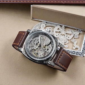Puede incluir: Un reloj esqueleto plateado con una correa de cuero marrón. La esfera revela el mecanismo interno. La correa está cosida con hilo marrón claro. El reloj se muestra sobre una superficie neutra. La correa mide unos 22 cm de largo.