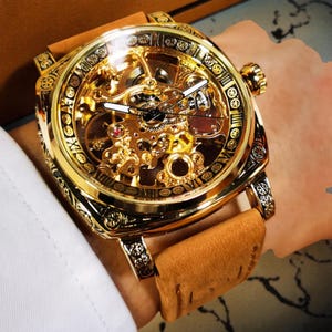 May include: A gold-coloured, skeleton wristwatch with a square face and a brown leather strap. The watch displays its intricate internal mechanisms through a clear face. It features Roman numeral hour markers and decorative detailing around the bezel.