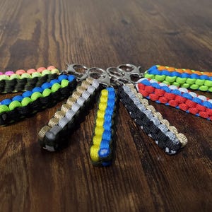 May include: A collection of colorful paracord keychains. Each keychain features a woven design in various color combinations, including black, blue, green, red, and yellow. The keychains are attached to silver-colored metal rings.