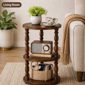 May include: A three-tiered round wooden side table with turned legs. The top shelf holds a speckled white planter with a green plant and a floral teacup. The middle shelf holds a rectangular radio. The bottom shelf holds a woven basket with a bear.