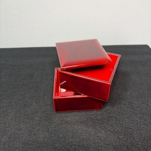 Könnte beinhalten: Eine glänzende, rote rechteckige Box mit Deckel, teilweise geöffnet. Die Box besteht aus einem glatten, reflektierenden Material, und der Deckel ist leicht geöffnet, wodurch das Innere sichtbar wird. Die Box befindet sich vor einem dunklen Hintergrund.