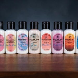 May include: A row of ten small body lotion bottles with black caps and white labels. Each label has a colorful design and the words "Buddah Milk Body Lotion". The bottles are arranged on a wooden surface.