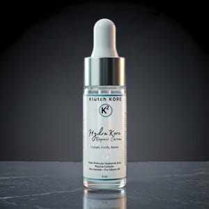 May include: A clear glass bottle of Klutch Kore Hydra Kore Repair Serum with a white dropper and silver cap. The label includes the brand name, product name, and key ingredients. The bottle contains 5 ml of serum.