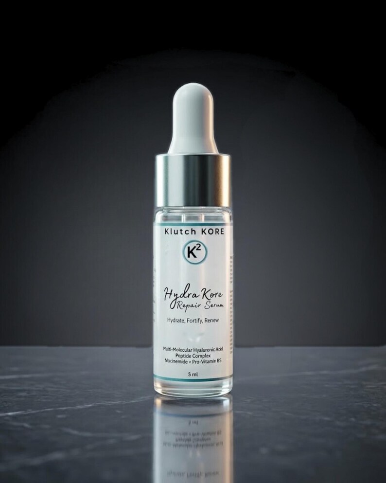 May include: A clear glass bottle of Klutch Kore Hydra Kore Repair Serum with a white dropper and silver cap. The label includes the brand name, product name, and key ingredients. The bottle contains 5 ml of serum.